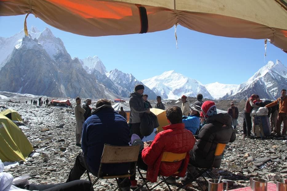 Trekkers seated under the flysheet of a dining tent at a rocky Concordia camp, tents and climbers around them and snow peaks behind