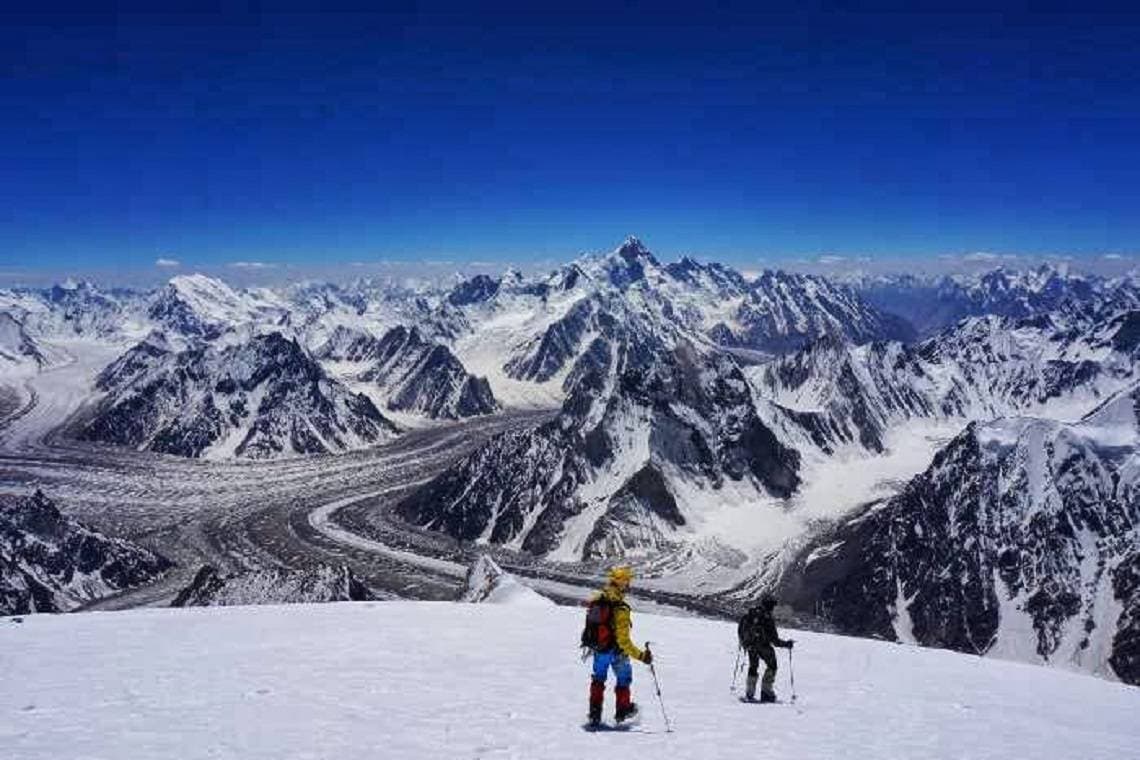 Two trekkers on a high snow slope above a wide glacier junction, a pyramid peak on the skyline