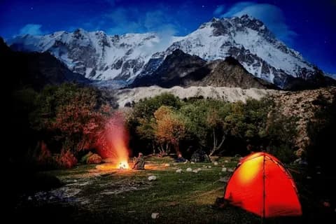 A red tent glowing beside a campfire on a grassy clearing with trees, snow peaks under a blue twilight sky