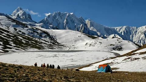 Trekkers and a blue tent beside a snow-covered lake basin with white peaks behind