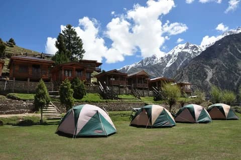 Four camping tents on a green lawn in front of wooden cabins with snow-covered peaks rising behind
