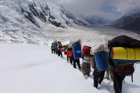 Porters carrying blue drums and loads in single file across a snowfield towards the Karakoram peaks