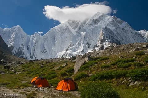 Two orange tents on a green alpine shelf beneath a huge snow-and-rock wall lit by the sun