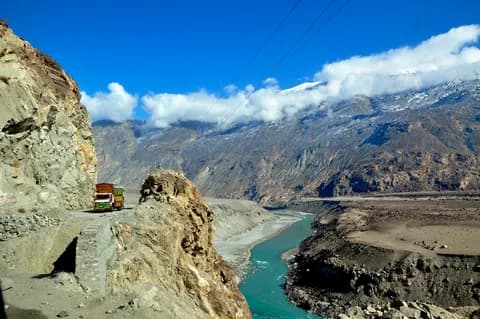 A colourful truck on the narrow cliff road above a turquoise river in a dry rocky gorge, snow peaks beyond