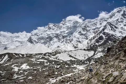 Trekker with a backpack on a rocky moraine path below a broad snow-covered mountain wall