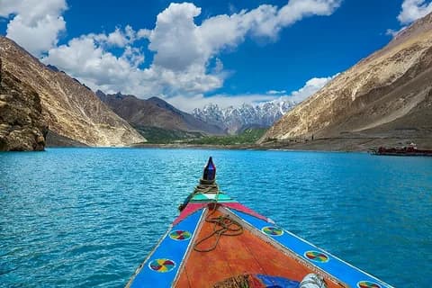 The bow of a wooden boat on a turquoise lake between steep golden slopes with snow peaks ahead
