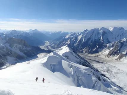 Two tiny figures on a snowy ridge high above a glacier valley ringed by white peaks