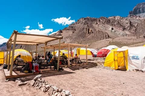 Dining shelter under a wooden frame with benches, yellow and red tents on stony ground beside an ochre rock face