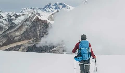 A trekker in a blue jacket and pack with trekking poles climbing towards cloud-covered snow slopes