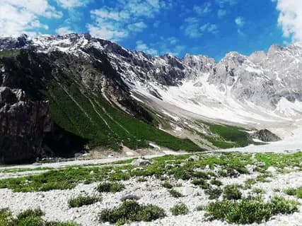 Green slope strewn with white boulders above a glacier, snow-covered Karakoram peaks and clouds behind