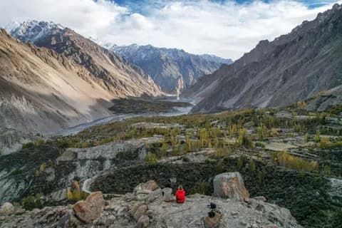 A walker in a red jacket looking out over the Hunza valley from a rocky ledge, autumn trees and snow peaks beyond