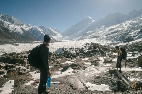 Two trekkers with packs on the moraine at the edge of a snow-covered glacier valley