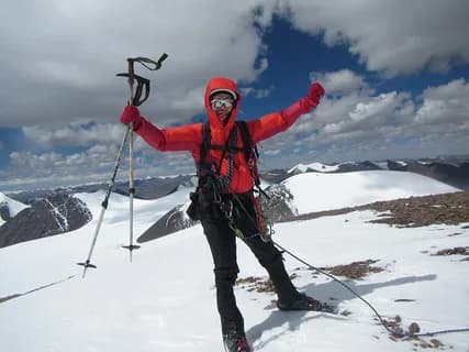 A climber in a red down suit raising both arms on a snow slope, rope and poles beside him and snowy peaks below