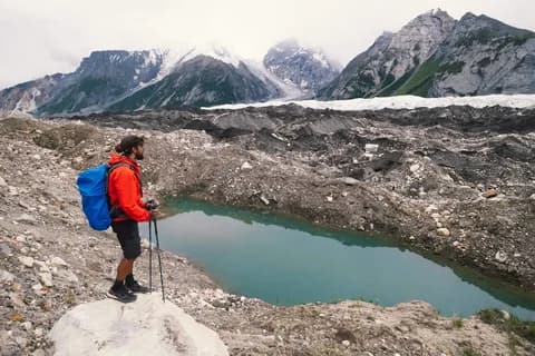 Trekker in an orange jacket standing above a turquoise meltwater pool among glacier moraine and ice