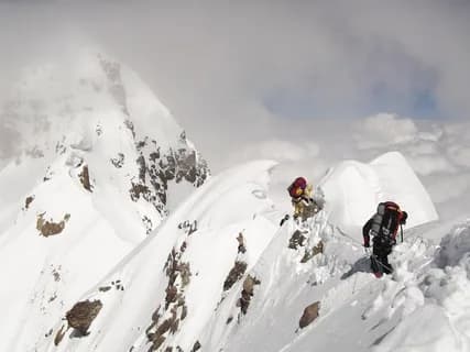 Two climbers with heavy packs ascending a steep snow ridge between rock outcrops and misty peaks