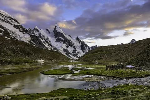 Braided streams and green marsh on a valley floor below a row of snow-covered rock spires under a clouded evening sky
