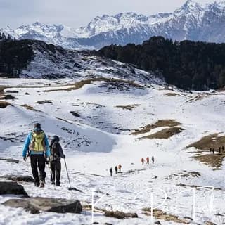 Trekkers with hiking poles descending a snow-covered plateau below a line of snowy peaks