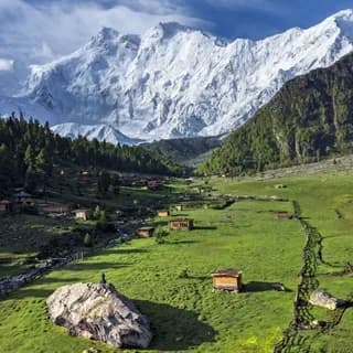 Wooden cabins scattered across a green meadow below a broad snow-covered mountain range