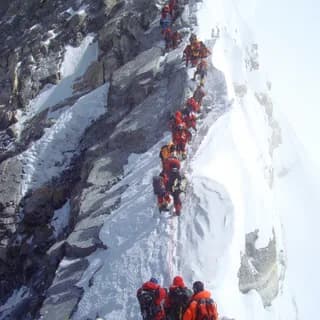 Line of climbers in colourful down suits ascending a fixed rope on a steep snow-and-rock face