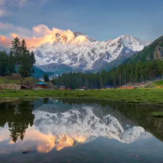 Snow-covered peaks lit by the setting sun reflected in a still lake ringed by pine trees