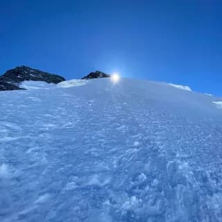 Sunlit snowfield rising towards a small rocky summit under a clear deep blue sky