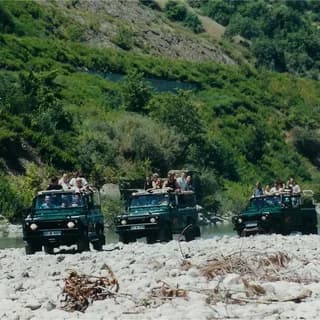 Three open safari jeeps carrying passengers parked on a stony riverbank below wooded hills