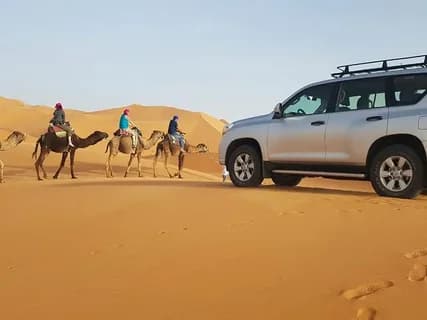 Camel riders and a 4x4 vehicle travelling across sand dunes