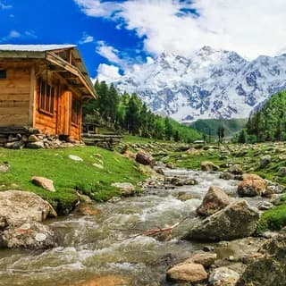 Wooden cabin beside a rocky stream below a snow-covered peak